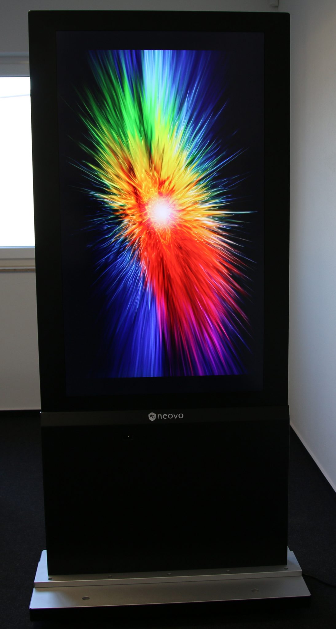 Samsung digital display screen showing a vibrant, multicolored burst of light with green, yellow, blue, red, and orange streaks radiating from a bright white center. The large LED screen stands upright in a modern indoor environment, highlighting its vivid color display and sleek design.