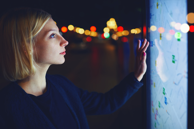 A woman with short blonde hair interacts with an illuminated digital map touchscreen at night, with blurred city lights in the background. The scene highlights modern navigation technology and urban exploration.
