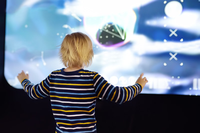 A young blond child in a striped shirt stands with arms raised, interacting with a large digital touchscreen featuring vibrant, colorful abstract shapes and patterns—demonstrating an interactive learning activity for kids.