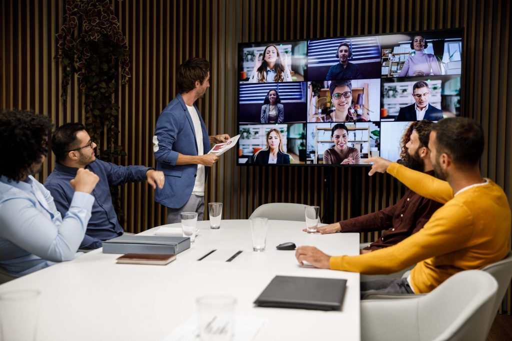 Team collaborating in a modern digital office conference room during a video call, with one person standing and pointing at a large screen showing remote colleagues, while others sit around the table and interact. Ideal for illustrating virtual meeting solutions or hybrid work environments.
