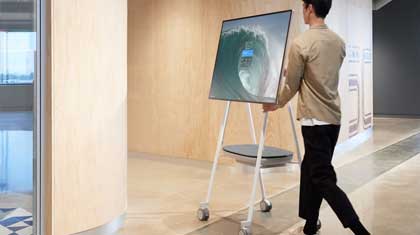 Person schiebt einen großen digitalen Bildschirm auf einen mobilen Standfuß durch eine moderne Büroflur mit hellen Wänden und glänzendem Boden. Der Screen, ideal für Kauf oder Miete, zeigt eine Wellen-Grafik.