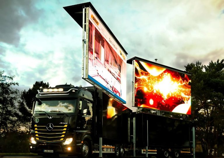 Schwarzer Mercedes-Truck mit zwei großen, erhöhten digitalen Werbetafeln, die leuchtende, bunte Grafiken anzeigen; der Truck steht im Freien vor bewölktem Himmel und Bäumen. Ideal zum Kaufen oder Mieten als mobiler LED-Werbeträger.