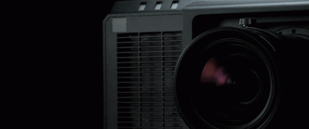 Schwarzer Beamer (Projektor) - Nahaufnahme eines eingeschalteten Projektors, dessen Linse hell leuchtet und einen Lichtstrahl nach vorne wirft, vor dunklem Hintergrund; ideal für alle, die einen Beamer kaufen oder mieten möchten.
