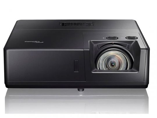 Optoma Beamer in Schwarz, Frontansicht: Der Projektor steht auf einer weißen, spiegelnden Oberfläche. Rechts vorne ist das große Objektiv deutlich sichtbar, oben befinden sich die Bedienknöpfe. Ideal für Heimkino und Präsentationen.